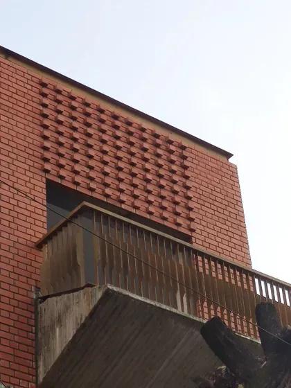 A corner detail showing the cantilevered balcony with its concrete base and simple railing, set against the patterned brick wall.