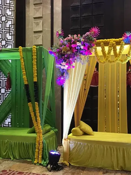 Individual seating pods for a Mehendi ceremony, draped in vibrant green and yellow fabrics and topped with colorful floral bouquets.