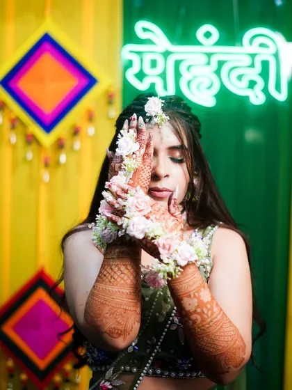 This artistic pose highlights the detailed mehndi work on the back of the hands and forearms, perfectly matching the bride's floral jewelry and festive attire.