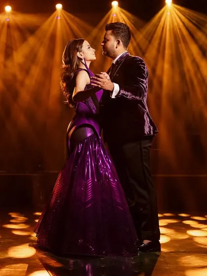 A dramatic shot from the masquerade ball, with the couple dancing under spotlights. The deep purple of their coordinated outfits creates a rich, cinematic feel.
