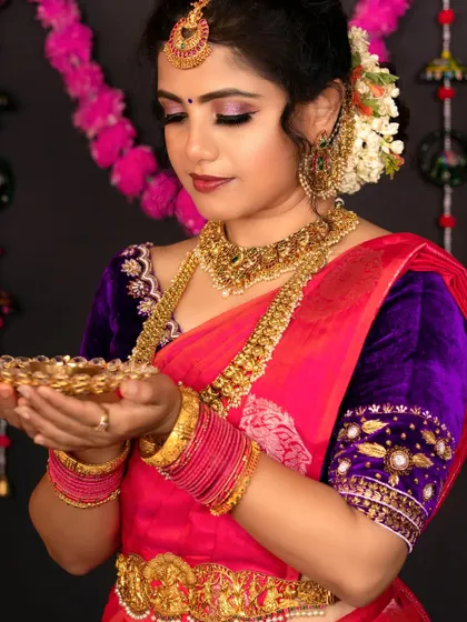A model in a pink and purple saree, holding a diya. She is wearing a grand temple jewellery set.
