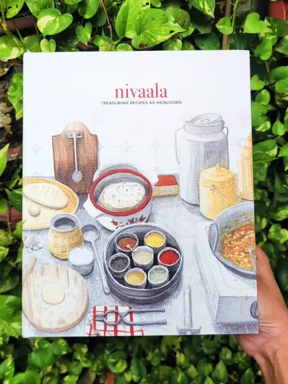 Our Nivaala journal, "Treasuring Recipes as Heirlooms," held against a backdrop of lush greenery. The cover, featuring artwork by Shawn D’Souza, is a celebration of the `ghar ka khana` we often take for granted. It's a stunning present for friends and family.