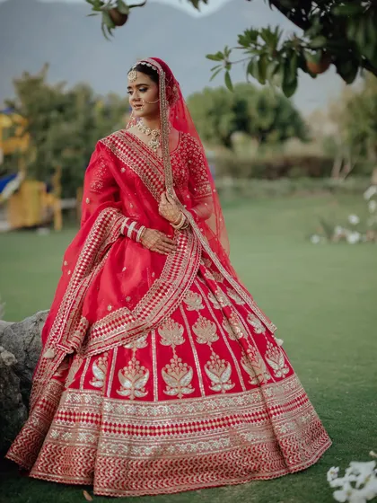 The bride in her red lehenga, captured in a beautiful garden setting, looking like a classic painting.