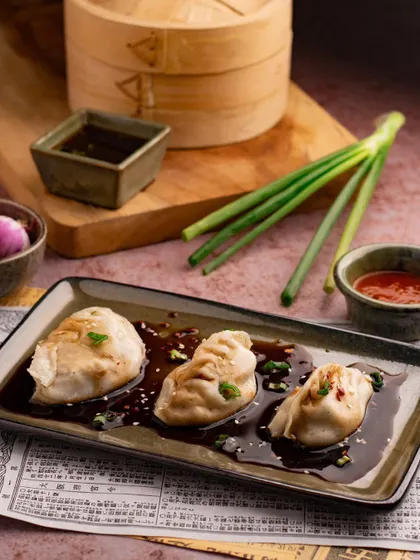Steamed dumplings in a savory sauce, styled with an Asian-inspired aesthetic. The use of a bamboo steamer and spring onions in the background adds authenticity and context to the shot.