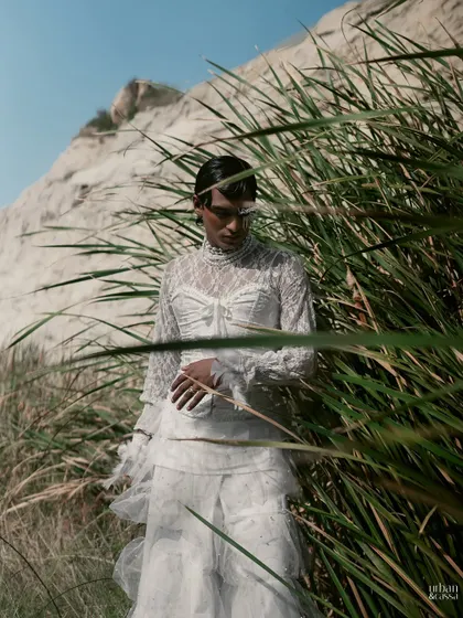 The model is framed by tall grass, adding a natural element that contrasts with the highly stylized makeup and hair.