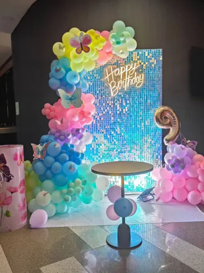 Another view of the vibrant butterfly theme decoration. This angle shows the blue sequin backdrop, the "Happy Birthday" neon sign, and the colorful balloon arrangement creating a festive photo corner.