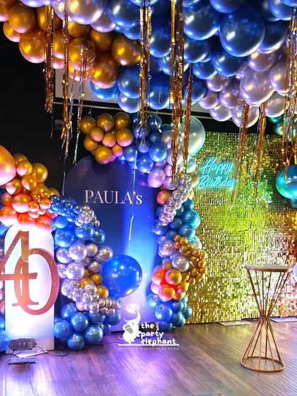 A personalized backdrop for Paula's 40th birthday, featuring her name, a neon sign, and an elaborate blue, gold, and silver balloon arrangement against a gold sequin wall for maximum sparkle.