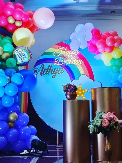 A vibrant and cheerful rainbow theme for a first birthday. The decor features multiple rainbow arches, colorful balloon clouds, and a personalized milestone board detailing the baby's first year.