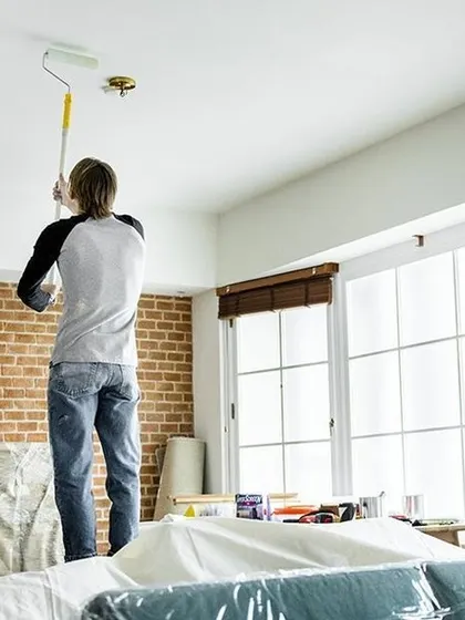 A painter applies a fresh coat of white paint to a ceiling using a roller with an extension pole. We use professional tools and techniques, like covering furniture with plastic sheets, to protect your home and deliver a clean job.