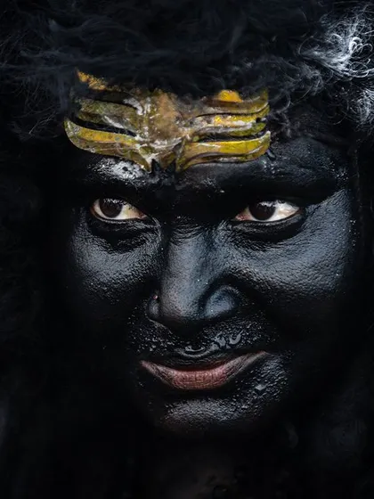 An extreme close-up of a Kulasai performer. The detailed face paint and direct, piercing gaze create an intimate and powerful portrait that feels both human and divine.