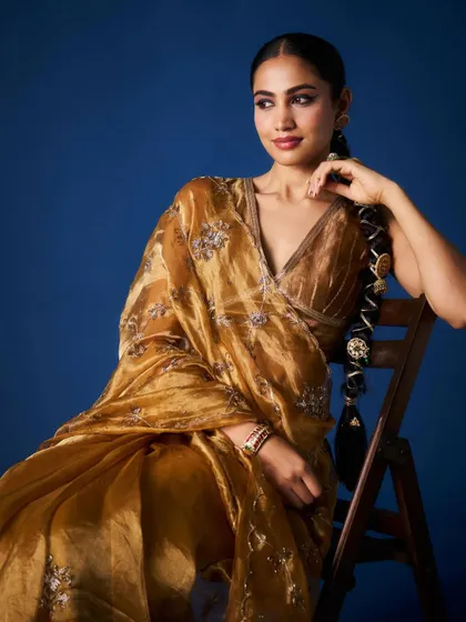 A seated portrait showcasing the beautiful fall of the golden organza saree. The styling is kept elegant and minimal to let the richness of the fabric and the intricate embroidery be the focus.