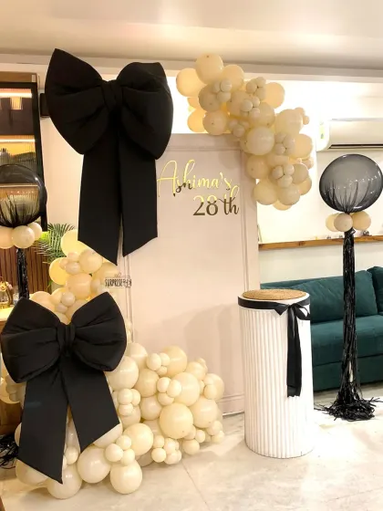 A closer look at the chic "bow" themed birthday decor. This setup includes a clean white backdrop with custom gold lettering, elegant black bows, and a stylish arrangement of cream-colored balloons for a sophisticated celebration.