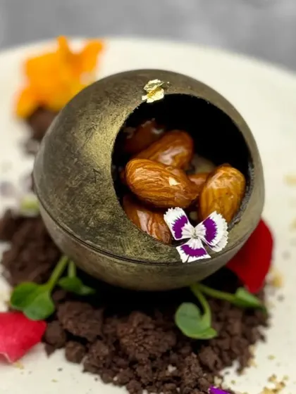 A close-up of our Ginger and Almond Chocolate Sphere. The golden-dusted chocolate shell, edible flowers, and rich chocolate soil create a dessert that is a true work of art.