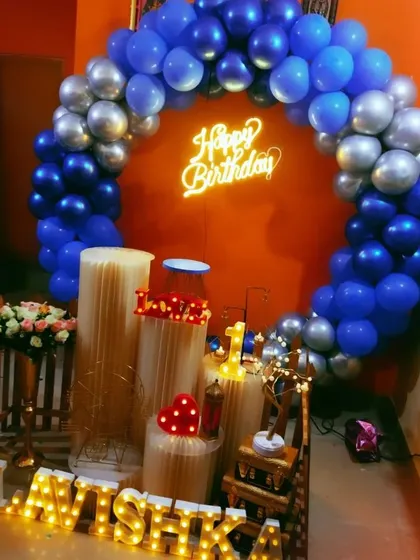 A beautiful home birthday decoration for Lavishka, featuring a blue and silver balloon ring against a vibrant orange wall.