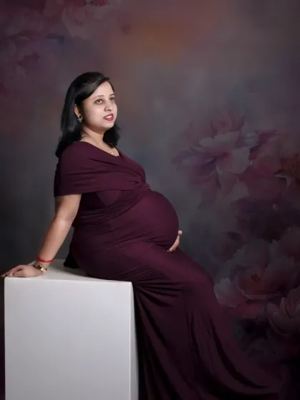 A classic and elegant solo portrait. The mom-to-be sits gracefully, her dark red gown flowing around her, against a soft, painterly backdrop.