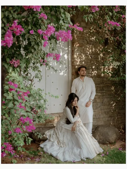 A stylish portrait of a couple against a stone wall adorned with vibrant pink bougainvillea. Their coordinated white outfits and poised expressions create a chic and modern pre-wedding photo.