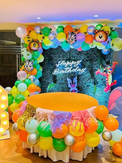 A jungle-themed birthday setup with a focus on the cake table. The table is decorated with colorful balloons and placed in front of a green foliage backdrop with a neon sign.