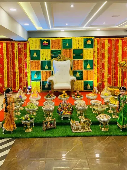 This traditional Seemantha decor features a checkered backdrop in auspicious green, yellow, and red. The entire setup, including the 21 plates and doll arrangements, is placed on a neat artificial grass carpet.