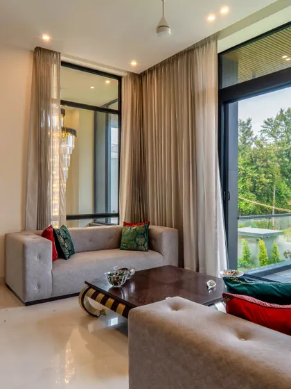 Another view of the farmhouse drawing lounge, showing the connection to the outdoors. The design uses large glass windows and sheer curtains to maintain an airy, open feel while ensuring comfort.