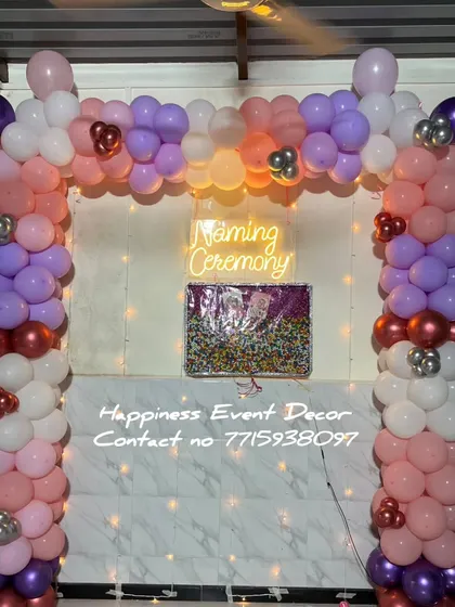 This balloon arch for a naming ceremony uses a beautiful color palette of pastel pink, purple, and white. The 'Naming Ceremony' neon sign and fairy lights in the background create a warm glow.