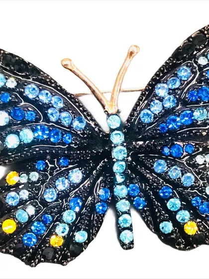 A close-up of our blue crystal butterfly brooch. The wings are detailed with various shades of blue and yellow crystals on a black metal frame, creating a beautiful mosaic effect.
