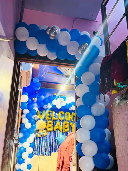 An entrance decoration for a 'Welcome Baby Boy' celebration. A blue and white balloon arch frames the doorway, leading into the decorated home.