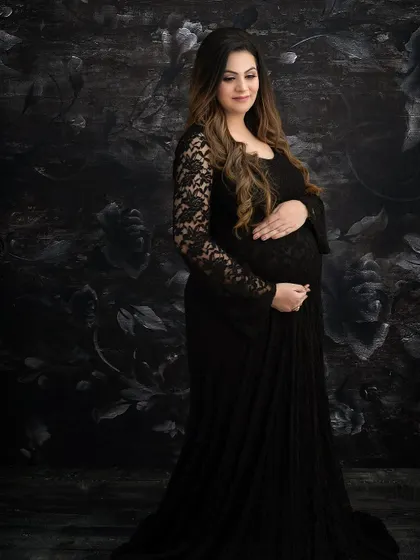 A classic maternity portrait in a black lace gown. The dark floral backdrop adds a touch of gothic romance and texture, resulting in a timeless and elegant image.