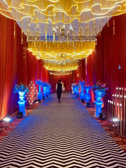 A modern and chic entrance with a black and white chevron carpet. The walkway is illuminated by blue uplighting on the vases and a unique wavy, lit ceiling treatment.