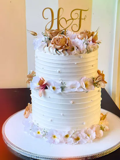 A different perspective of the elegant two-tier wedding cake, showing the beautiful initial topper and the careful placement of each flower. This cake is made with premium cream cheese frosting and white chocolate.