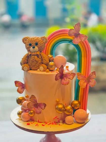 A cute and modern teddy bear cake. The design features a sweet teddy bear sitting under a rainbow, surrounded by butterflies and pastel-colored spheres.