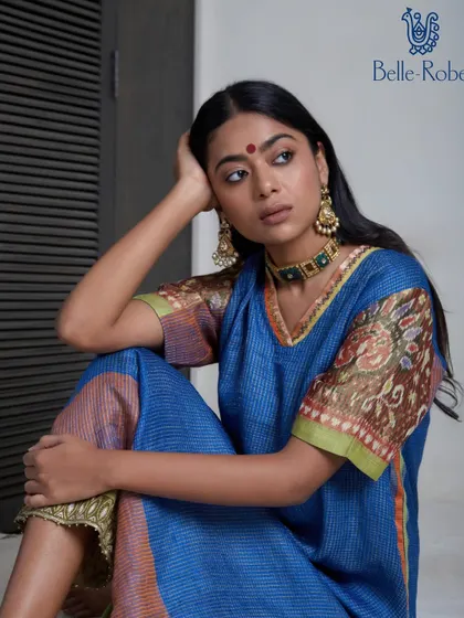 A close-up portrait for Belle Robe. The makeup is soft and traditional, with a bindi and subtly defined eyes, complementing the blue printed kurta and gold earrings.
