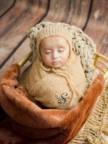 A newborn snugly wrapped in a beige knit swaddle and bonnet. The rustic wooden background and soft blankets create a warm, earthy, and cozy feel.