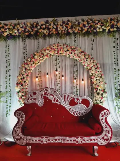 A vibrant reception stage featuring a stunning red velvet sofa with silver carving. The backdrop includes a full floral arch made of roses and chrysanthemums, hanging Edison bulbs, and an overhead floral runner, all set against a clean white drape.