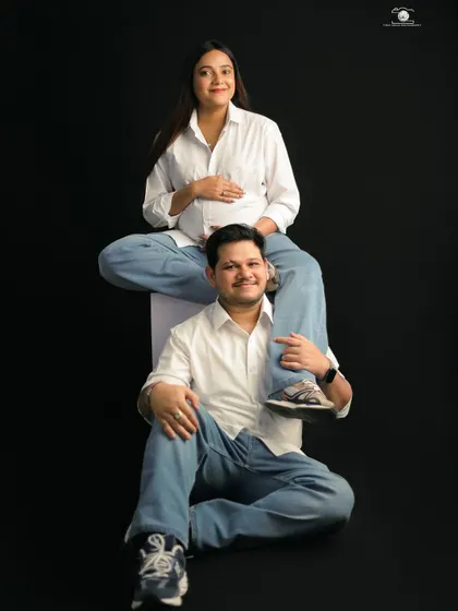 A playful and unique pose showing the couple's fun-loving personality. The mother-to-be sits on her partner's shoulders, both smiling at the camera in matching casual outfits.