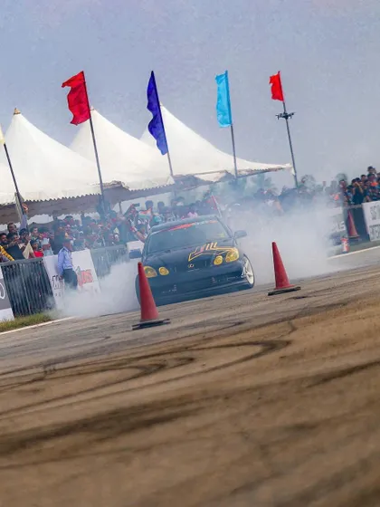 A drift car slides past the crowd, flags waving in the background. The combination of speed, smoke, and sound creates an electric atmosphere.