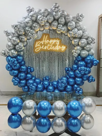 A cool-toned birthday decoration with a circular balloon frame in blue and silver. The setup includes a silver shimmer curtain and a neon sign, with balloons reflected on a glass table.