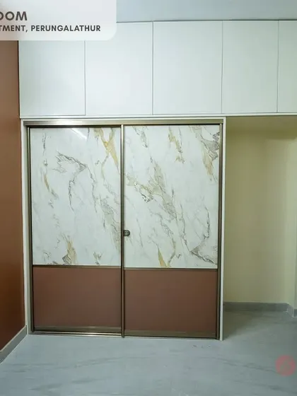 The second bedroom in the Perungalathur project showcases a stylish sliding wardrobe with a mix of marble-patterned and brown panels, complemented by white overhead storage units.