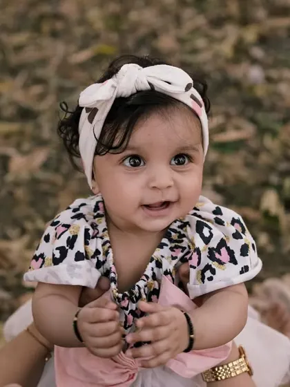 A close-up of a baby girl with a sweet smile, held up during an outdoor shoot. The natural background adds warmth to her happy expression.