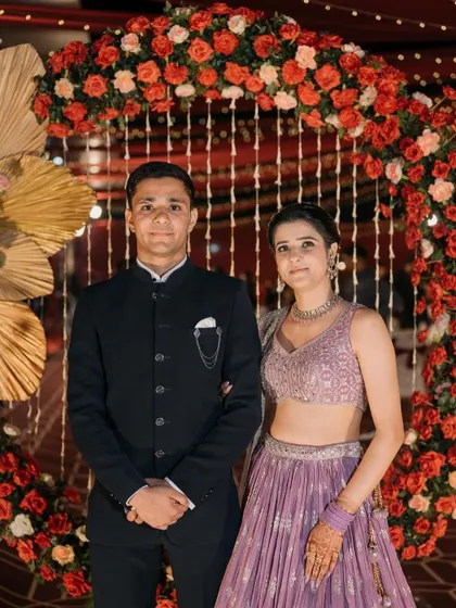 A happy couple posing for a photo against a stunning reception backdrop. The circular arch is decorated with red and orange flowers and accented with golden palm leaves, creating a warm and celebratory look.