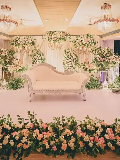 We design elegant and classic stage decor for receptions and engagements. This setup features a beautiful white chaise lounge against a backdrop of lush white and pastel pink floral arrangements, creating a sophisticated and romantic atmosphere.