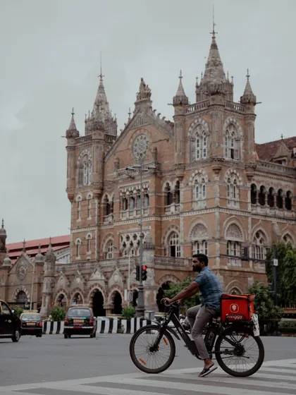 A delivery person on a bicycle crosses the road in front of the magnificent Chhatrapati Shivaji Maharaj Terminus (CSMT). This image juxtaposes the city's grand history with its modern-day hustle.