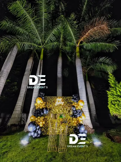 A surprise party setup featuring a gold shimmer wall nestled among palm trees, creating a glamorous and unexpected focal point for an outdoor celebration.