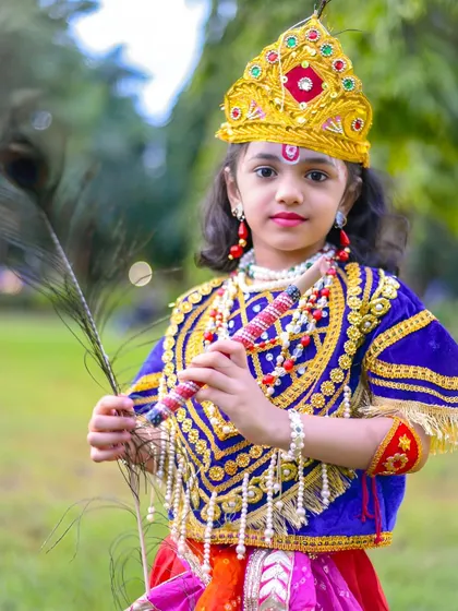 A beautiful close-up portrait of a young girl dressed as Krishna, holding a flute and peacock feather. The ornate crown and jewelry complete the look.