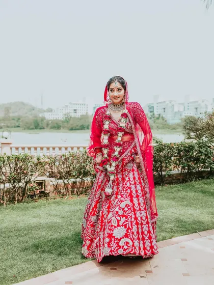 A full-length portrait of the bride by the lakeside, looking happy and beautiful in her red wedding lehenga.