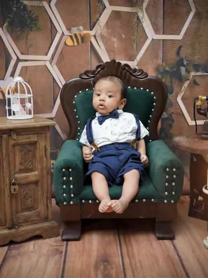 A portrait from a three-month milestone session. The baby is dressed in a smart bow tie and suspenders, sitting confidently in a green armchair.