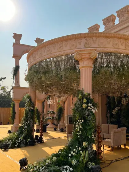 The sun shines on the magnificent Roman-style mandap, highlighting the architectural details and the lush floral arrangements that adorn it.