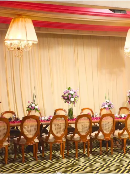 A long shot of an elegantly set dining table, ready for guests. The combination of classic chairs, floral centerpieces, and sophisticated lighting creates a truly royal ambiance.