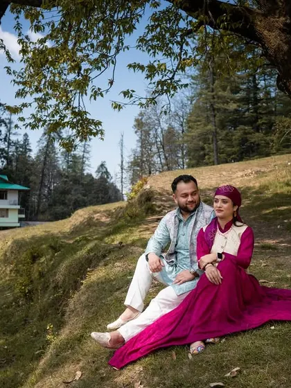 A wide shot showing the couple relaxing on a hillside, with a traditional Himachali house in the background, fully immersing them in the local culture.
