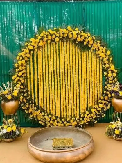 An outdoor Haldi setup with a circular marigold garland backdrop against a green screen. The arrangement includes a traditional brass 'urli' and matching floral arrangements on stands.