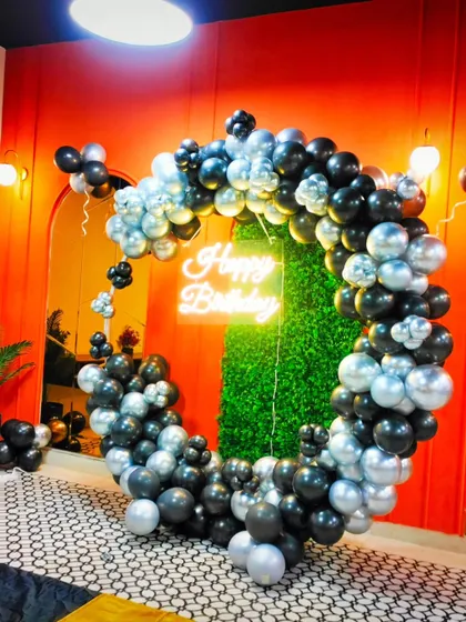 A bold and stylish ring decoration with black and silver balloons against a vibrant orange wall and a green hedge backdrop. The neon sign adds a cool, modern touch.